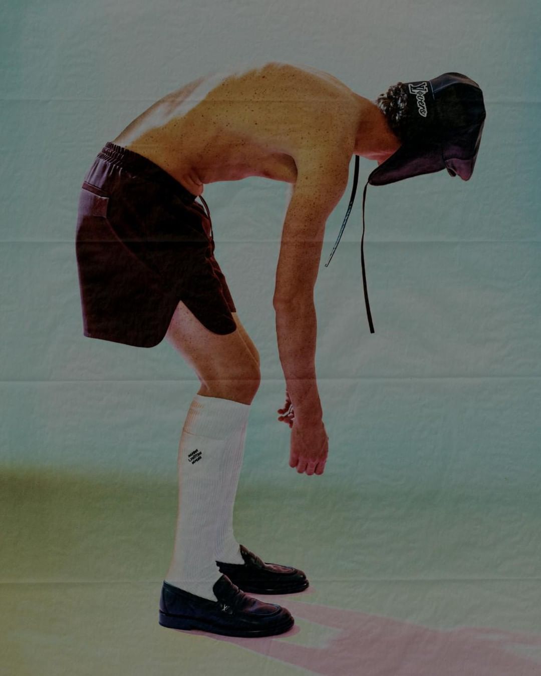 A shirtless person with a dark hat, shorts, socks and loafers is bent forward in a studio with a blue and pink tinted background.