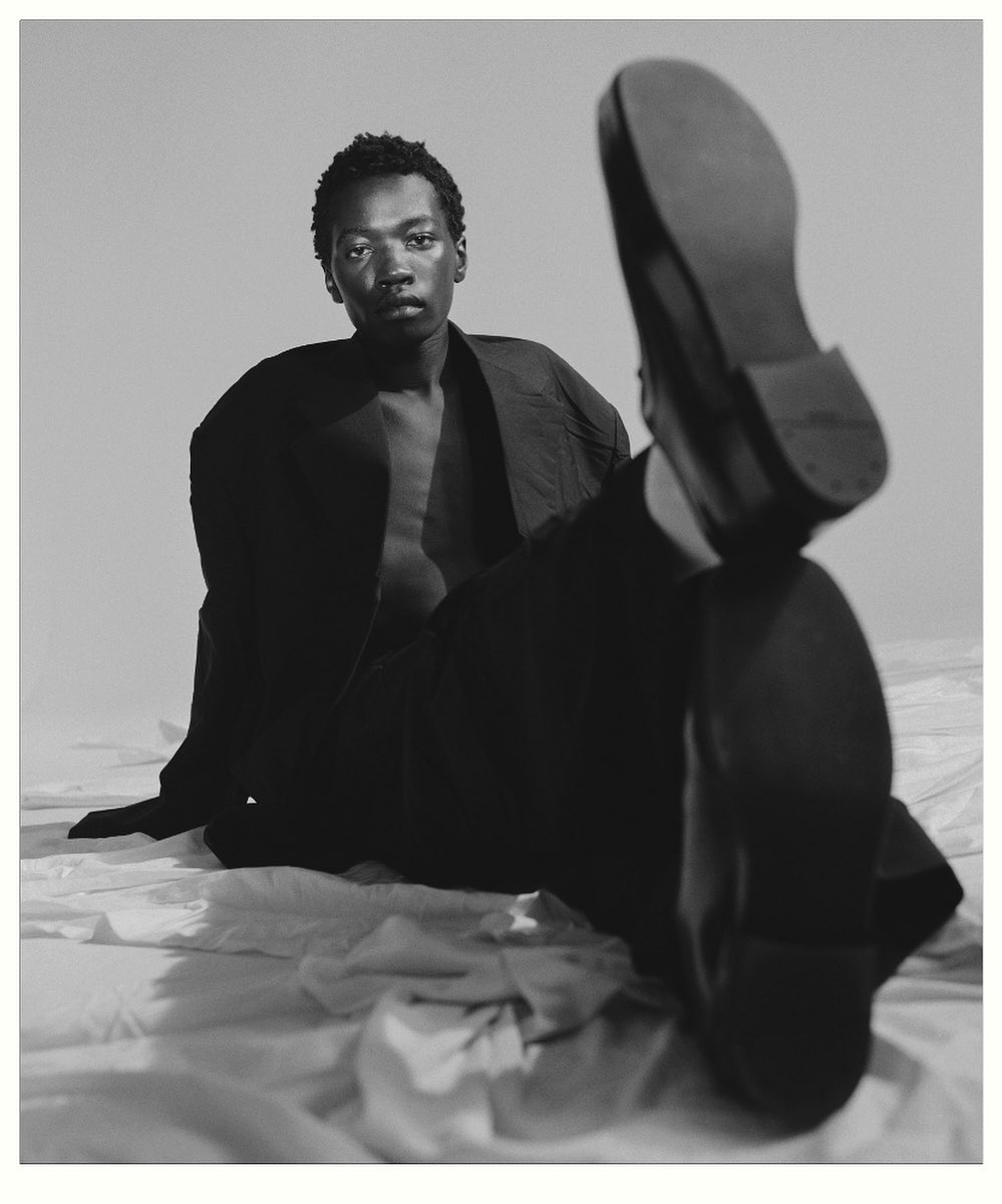 Black man seated on rumpled white fabric with one leg raised toward camera showing shoe sole