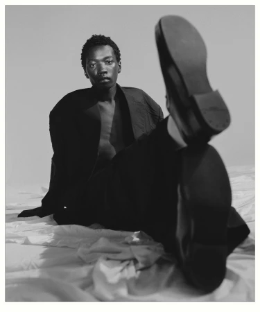 Black man seated on rumpled white fabric with one leg raised toward camera showing shoe sole