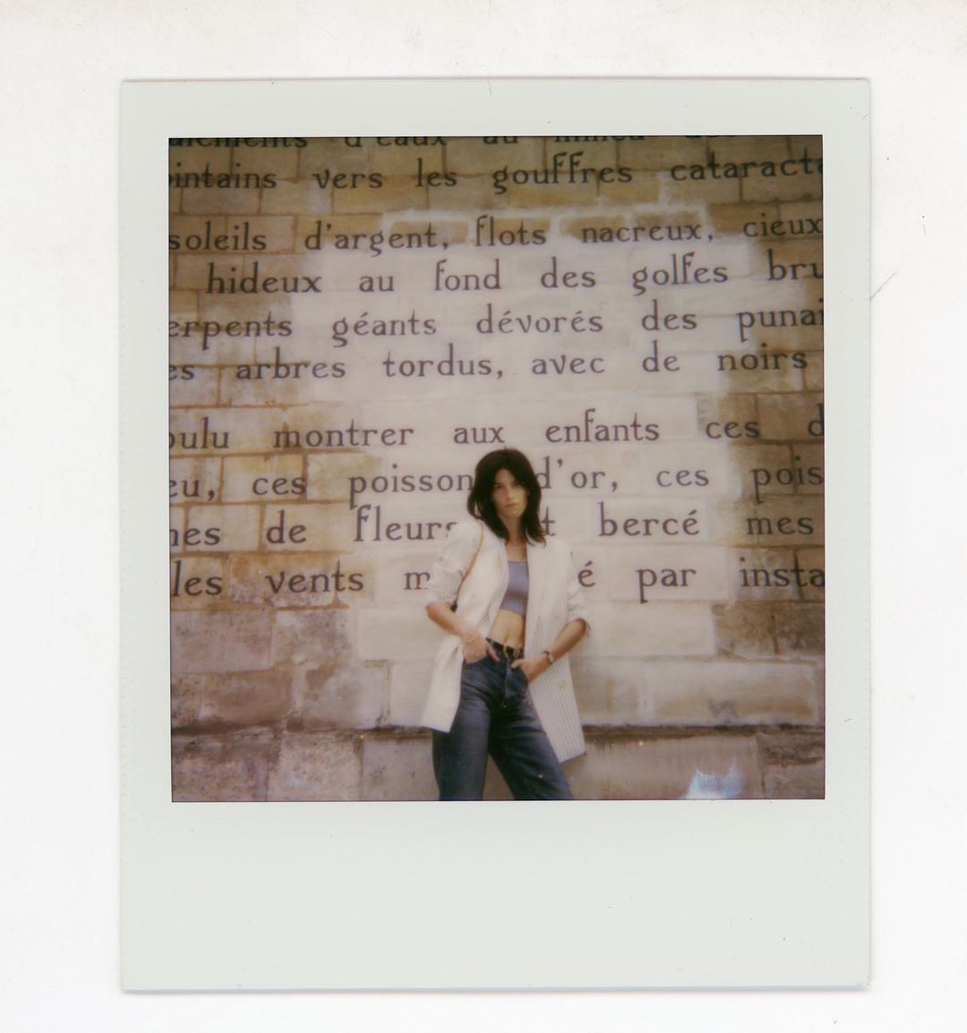 A young woman with dark hair poses against a vintage brick wall with large printed text in an indoor, editorial-style fashion shot.