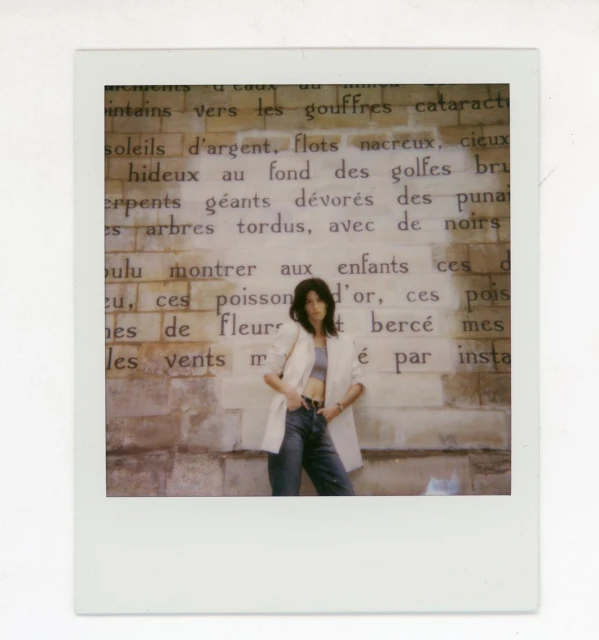 A young woman with dark hair poses against a vintage brick wall with large printed text in an indoor, editorial-style fashion shot.