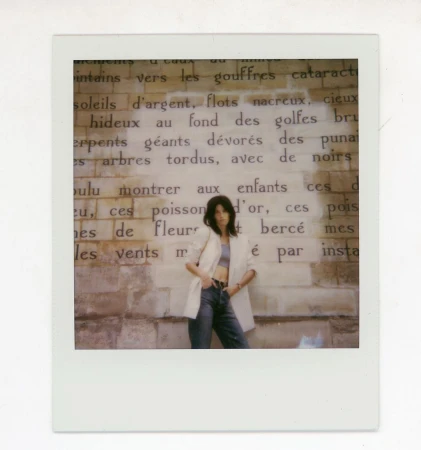 A young woman with dark hair poses against a vintage brick wall with large printed text in an indoor, editorial-style fashion shot.