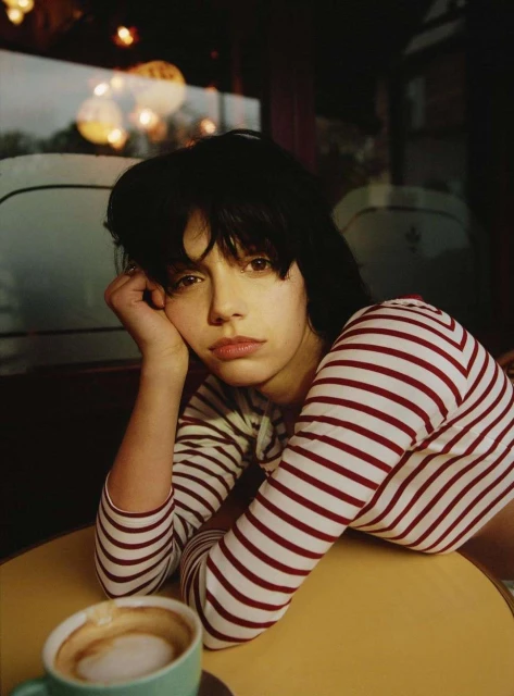 Indoor portrait of a young brunette woman with striped shirt resting at table with coffee in a cafe with soft, warm lighting.