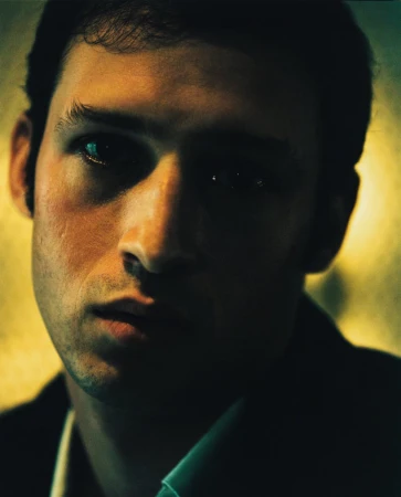 Portrait of a young adult male with dark hair and a collared shirt, looking at the camera with a serious expression in a dimly lit setting.