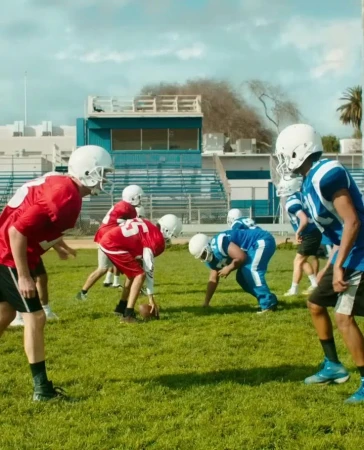 American football players in blue and red uniforms on a grassy field prepare for a play on a sunny day with a blue sky.