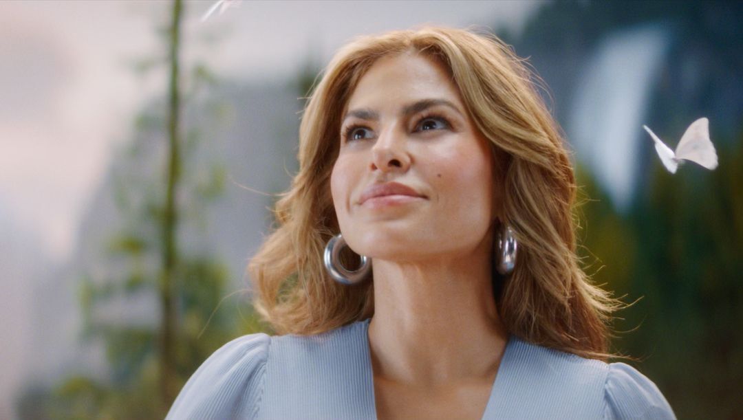 Woman with blonde hair poses in front of an outdoor scene with butterflies. She wears large hoop earrings and a light blue top.
