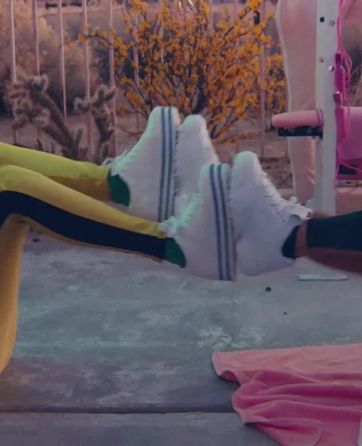 Two people doing leg extensions outdoors in white sneakers, yellow and black leggings, and pink sportswear with yellow flowers.