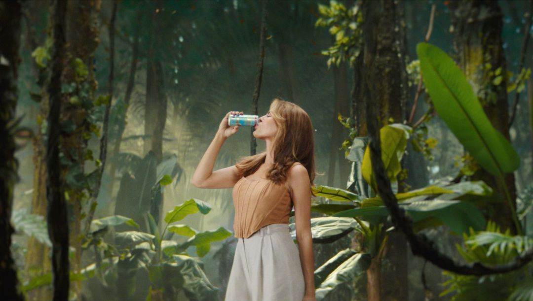 Adult woman drinks from a can in a verdant forest, wearing a tan top and white pants, surrounded by lush green foliage.
