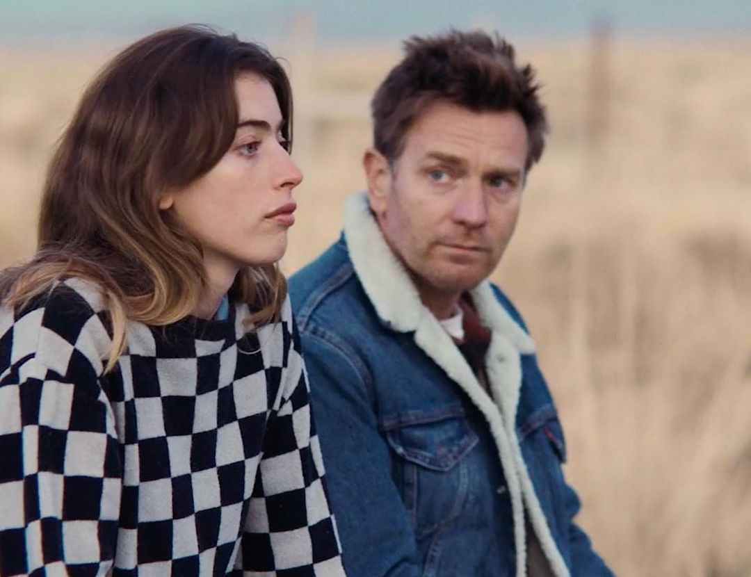 Two people, an adult woman in a checkered shirt and an adult man in a denim jacket, are outdoors with a muted, tan-colored background.
