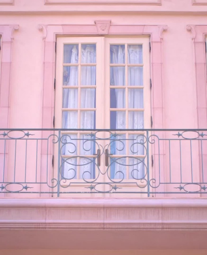 A soft pink building facade with a balcony featuring wrought iron railings, and a pair of wooden doors with glass panes and white curtains.