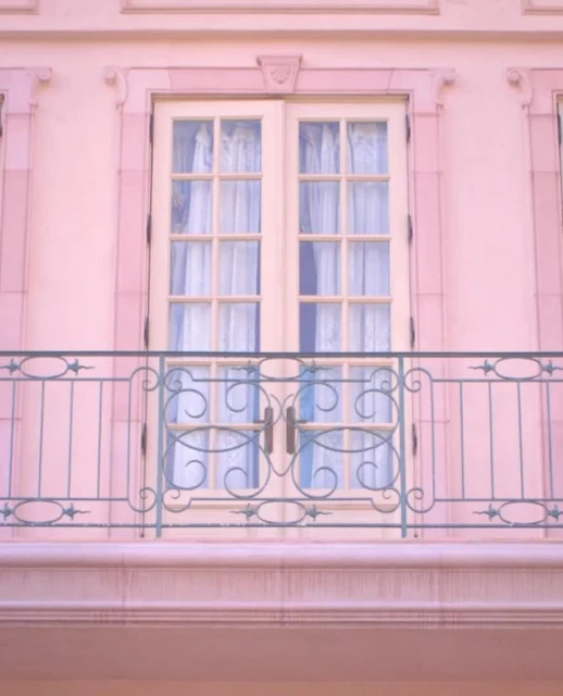 A soft pink building facade with a balcony featuring wrought iron railings, and a pair of wooden doors with glass panes and white curtains.