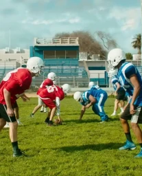 Football players in red and blue uniforms line up on a grassy field with stadium seating visible in the background.