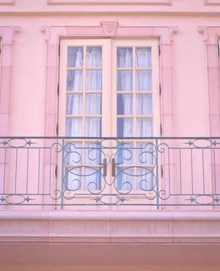 Close-up of double glass doors with white sheer curtains behind a decorative wrought iron balcony railing on a pink building exterior.