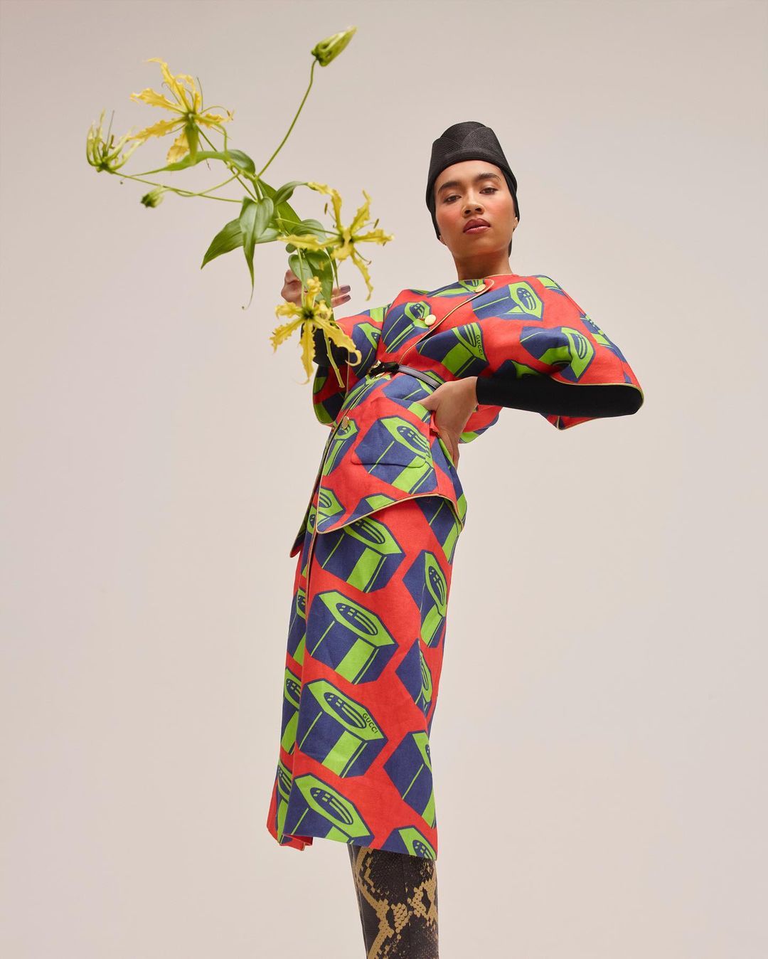 Adult female in a printed red kimono-style outfit poses with yellow tropical flowers against a neutral background in a studio shot.