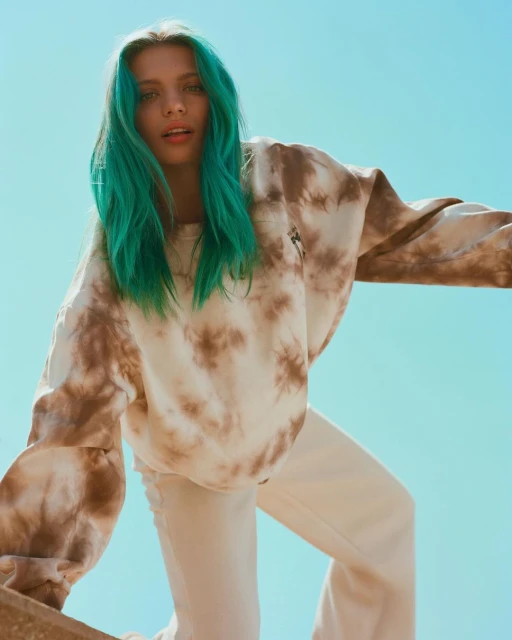 Young woman with turquoise hair poses wearing a tie-dye sweatshirt and cream pants against a clear blue sky in an outdoor setting.
