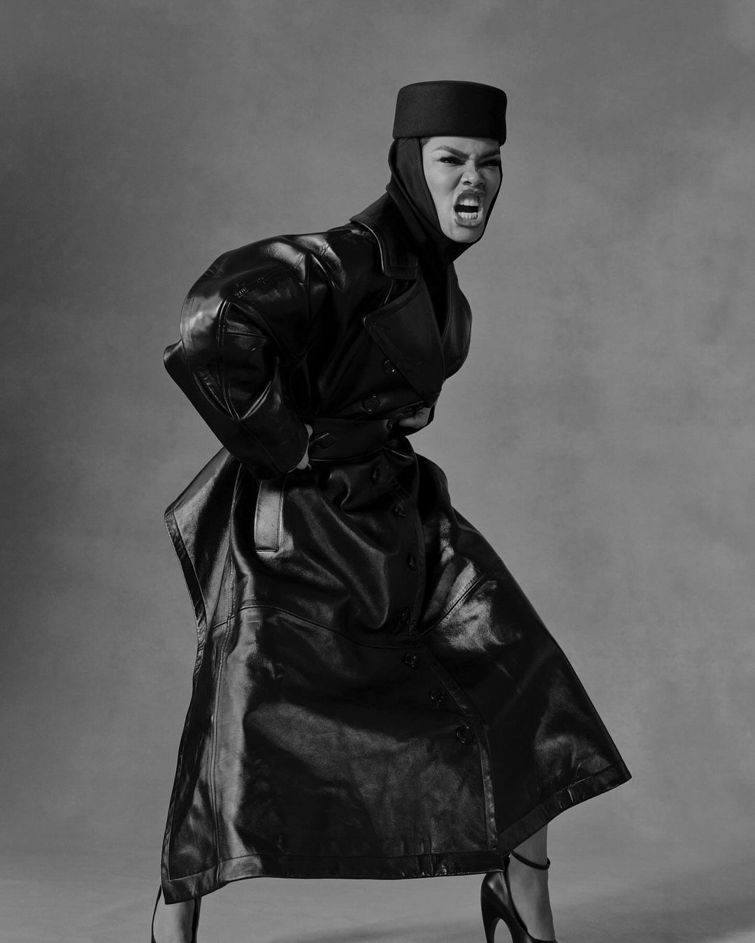 Monochrome portrait of a person in a black leather coat and hat, yelling and posing against a grey studio backdrop, high heels visible.