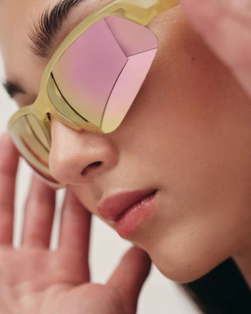 Close-up of a woman with a light skin tone wearing translucent yellow sunglasses with pink reflective lenses in a studio setting.