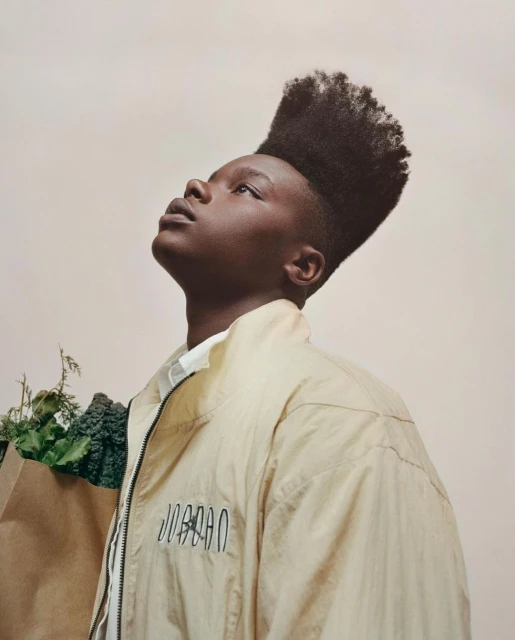 Portrait of a young adult male looking upward, wearing a jacket and holding a paper bag with green leafy vegetables.