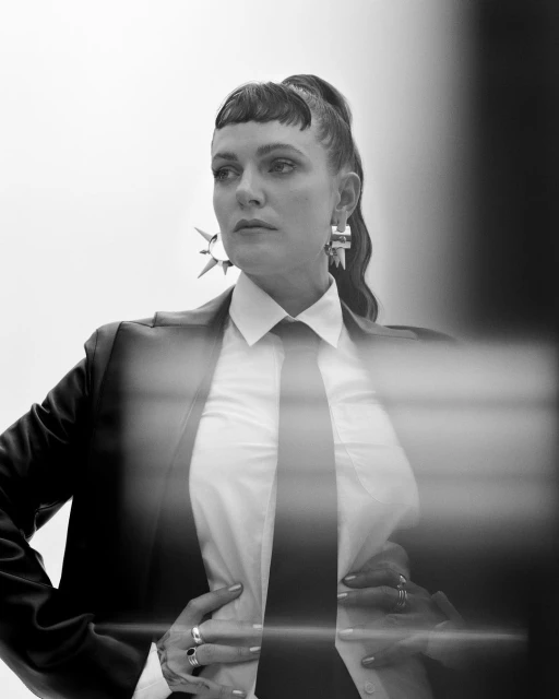 Monochrome portrait of a stylish adult woman with dark hair in a ponytail, wearing a suit jacket, tie, earrings and multiple rings.