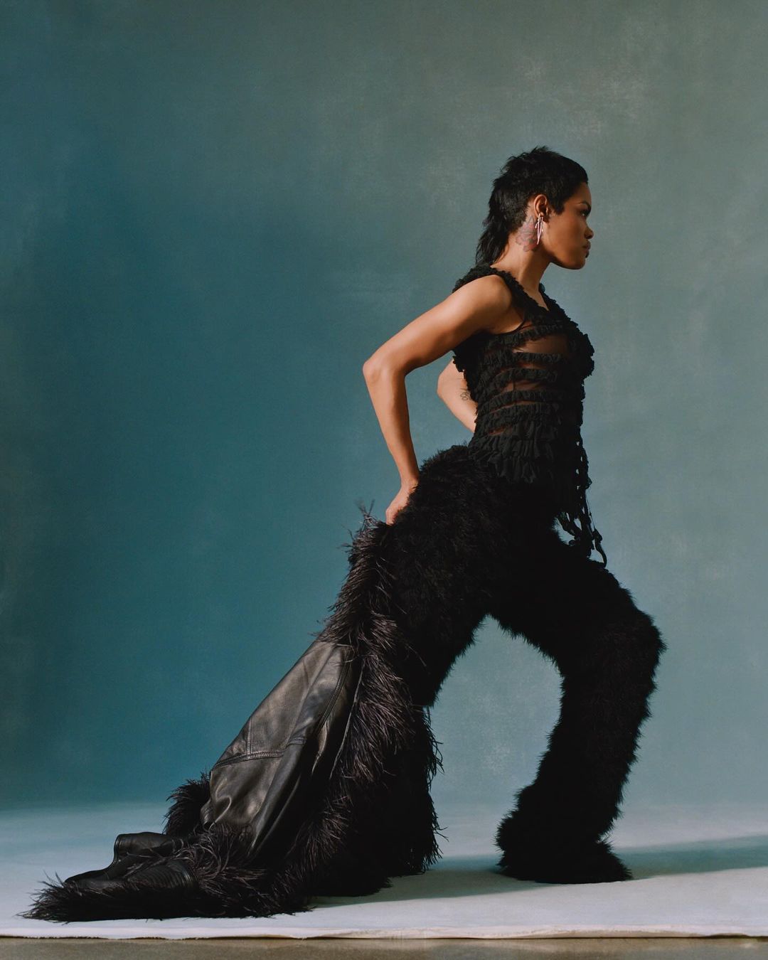 Woman in black feather-trimmed outfit strikes dynamic pose against muted teal backdrop in studio setting, side view, high-fashion editorial.