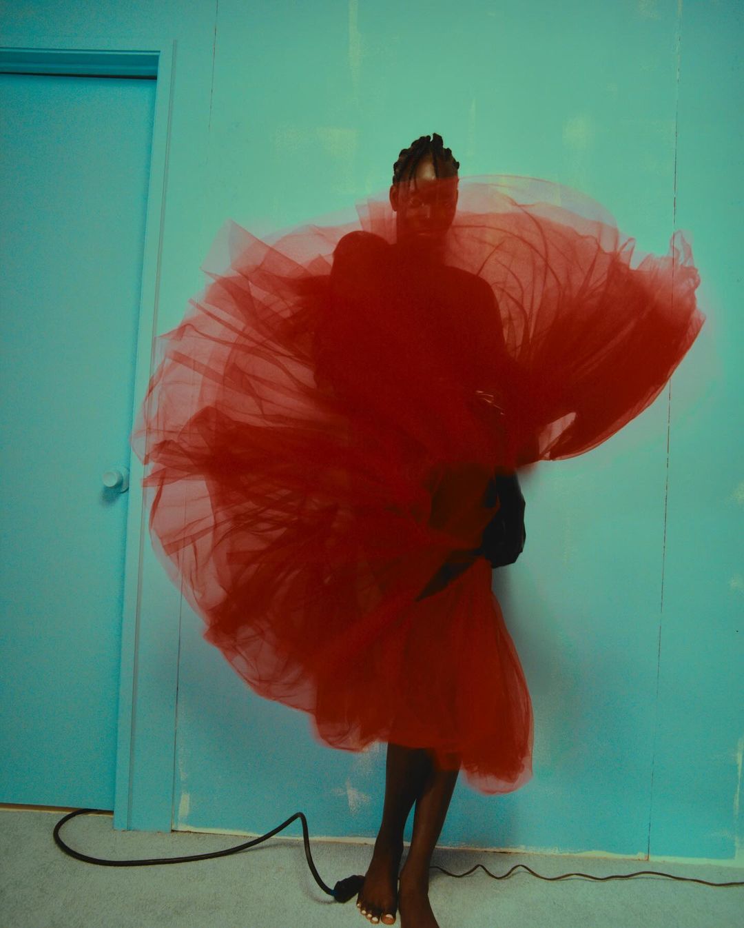 Woman in dramatic red tulle dress, barefoot, poses in a minimalist studio with a vibrant blue wall, creating a striking fashion portrait.