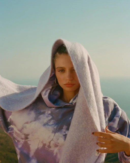 Young woman with light skin and neutral expression poses in front of a light blue sky, wearing a tie dye hoodie and hoop earrings.