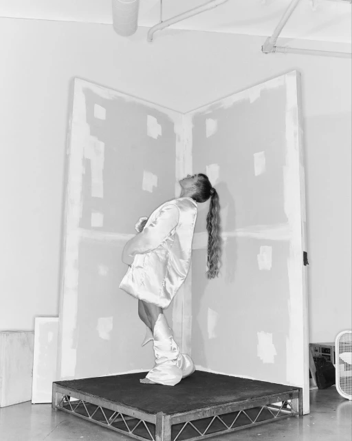 Woman in silky pajama set poses on a small platform against a makeshift drywall corner in a minimalist photo studio.