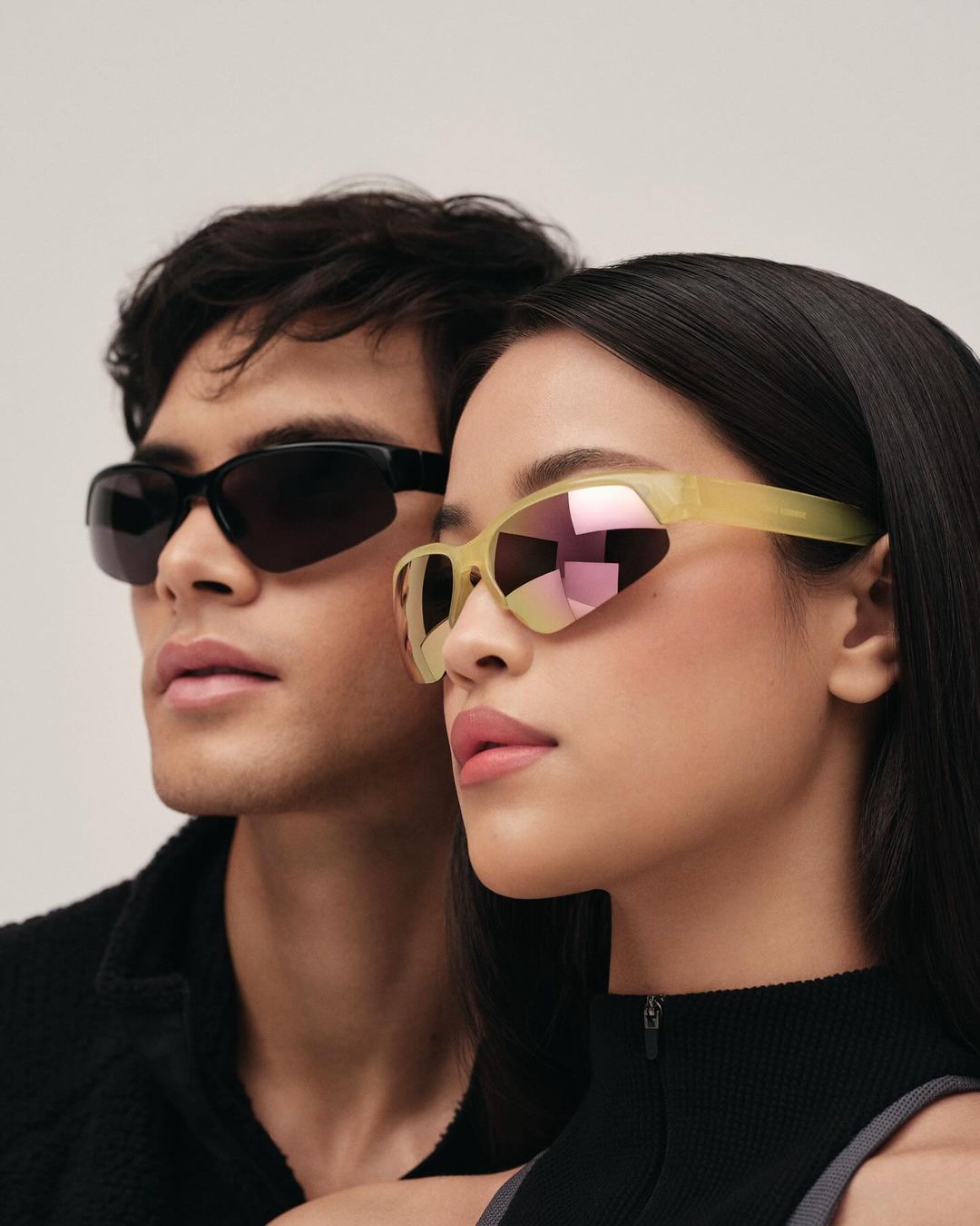 An attractive couple poses against a plain background while showcasing stylish, modern eyewear. The pair wear fashionable dark tops.
