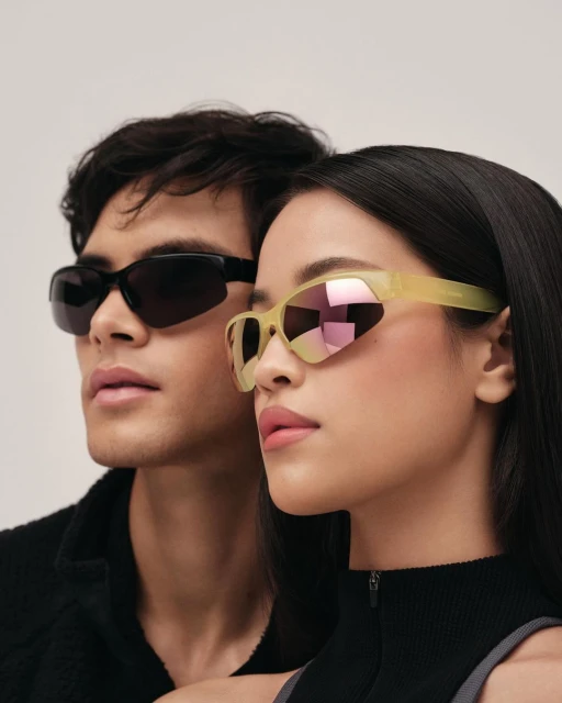 An attractive couple poses against a plain background while showcasing stylish, modern eyewear. The pair wear fashionable dark tops.