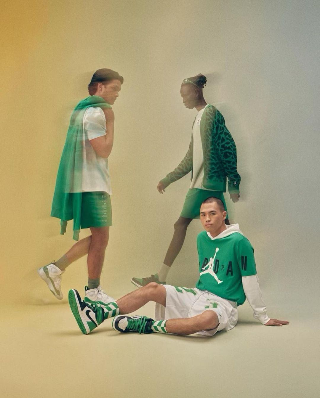 Three male fashion models showcasing green and white Jordan apparel in a studio setting with soft lighting and a gradient backdrop.
