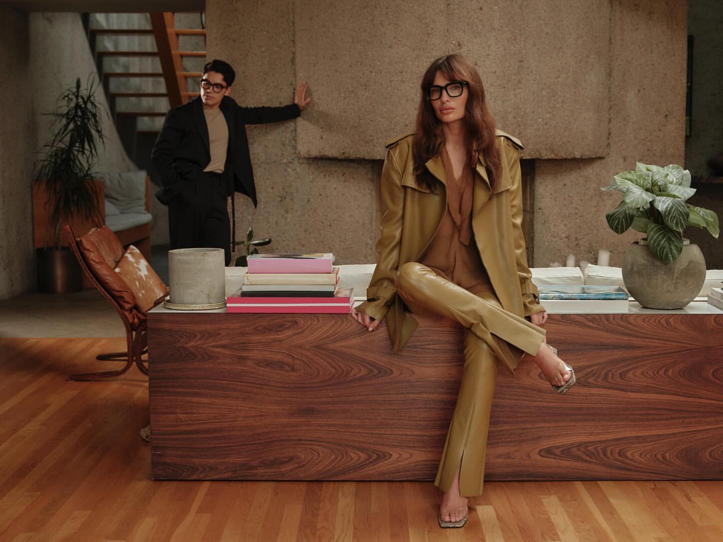 Two fashion models pose indoors. A woman in a tan trench coat and matching pants sits on a wooden surface. A man stands in the background.
