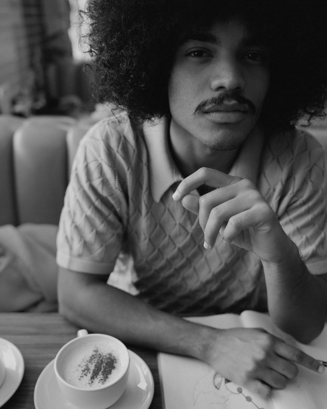 Black and white portrait of a man with an afro hairstyle sitting at a table with a cappuccino, drawing or writing in a notebook.