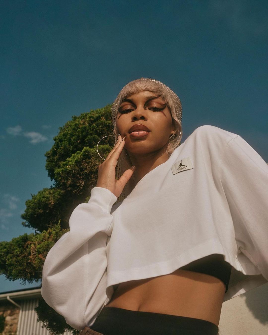 Woman with tan skin poses in a cropped white sweatshirt, black pants, and large hoop earrings in front of a tree and blue sky.