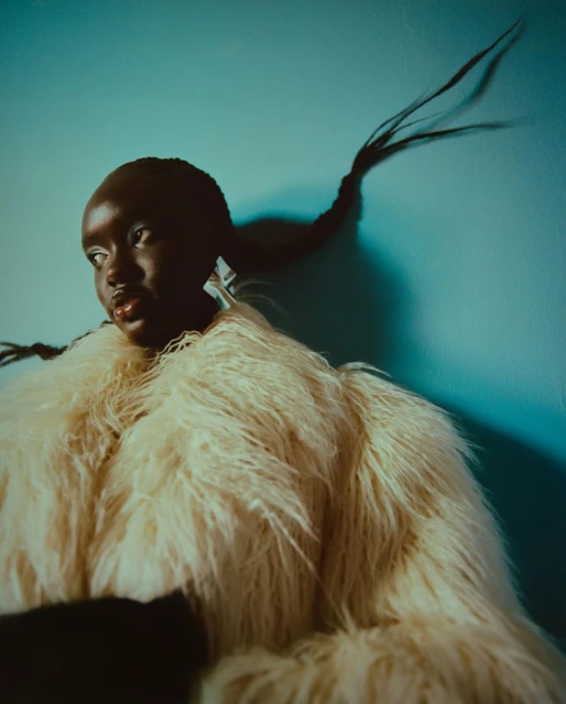Fashion shot of Black woman in faux fur coat posing against a teal backdrop with dramatic lighting highlighting her braided hair.