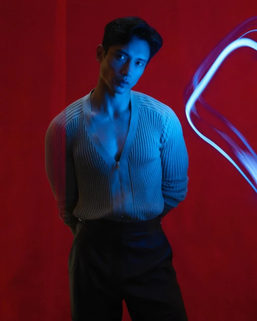 Portrait of an Asian man with dark hair, wearing a light-colored knit cardigan and dark pants, posing against a vivid red background.