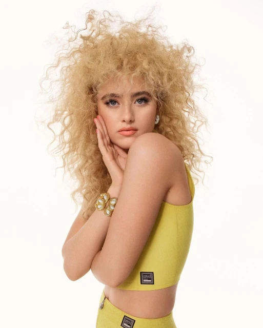 Blonde female model poses in a chartreuse crop top and skirt, hands near her face in a studio setting with a white background.