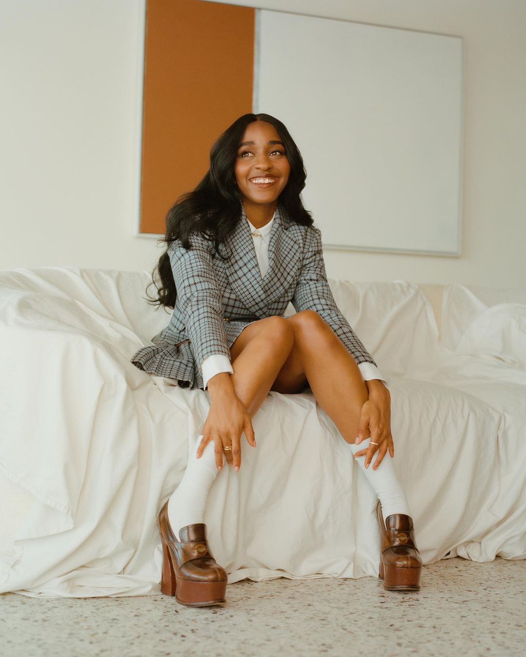 Stylish woman poses on a white couch in a grey plaid skirt suit with platform shoes. Indoor shot with a minimalist backdrop.