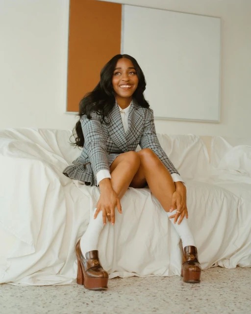 Stylish woman poses on a white couch in a grey plaid skirt suit with platform shoes. Indoor shot with a minimalist backdrop.