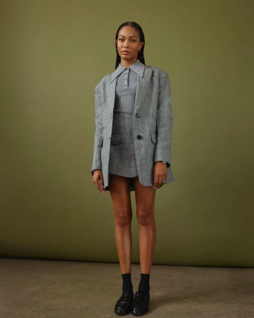 Woman posing in a gray tweed suit with a collared shirt and black loafers in a full shot against an olive green backdrop.