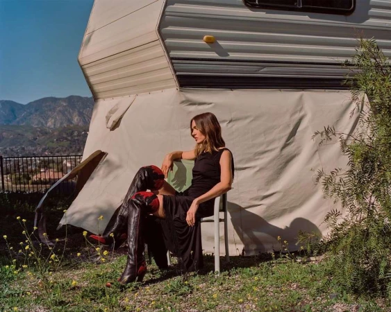 Woman in a black dress and tall red boots seated outdoors in front of a camper van by Emman Montalvan