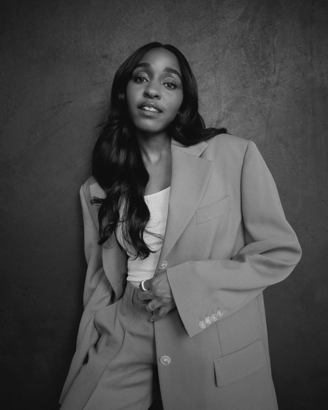 A young adult woman with long dark hair poses in a light suit against a mottled gray background in a studio portrait. She smiles softly.