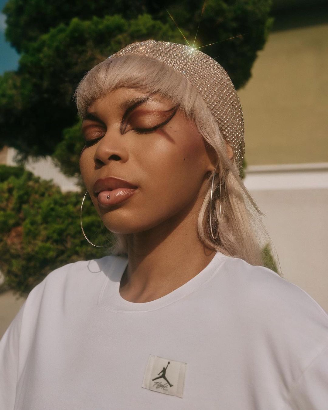 Close-up of a woman with closed eyes wearing jewelry headwear and a white T-shirt in a garden setting with green foliage.