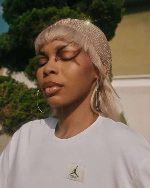 Close-up of a woman with closed eyes wearing jewelry headwear and a white T-shirt in a garden setting with green foliage.