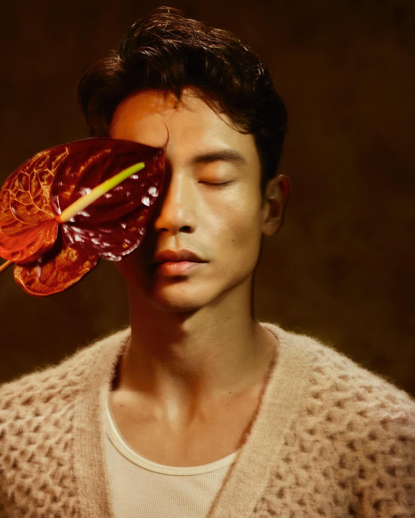 Close-up portrait of an adult male with dark hair and closed eyes, holding a red Anthurium flower, against a dark, blurred background.