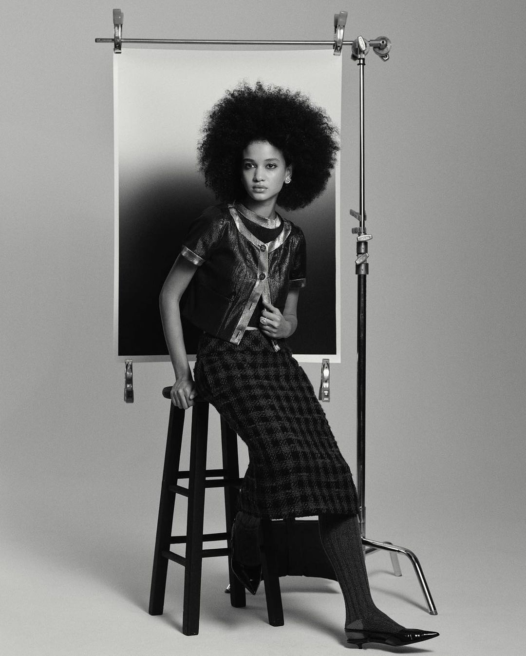 A woman with a large afro poses in a studio wearing a jacket and plaid skirt, sitting on a wooden stool with a gradient backdrop.