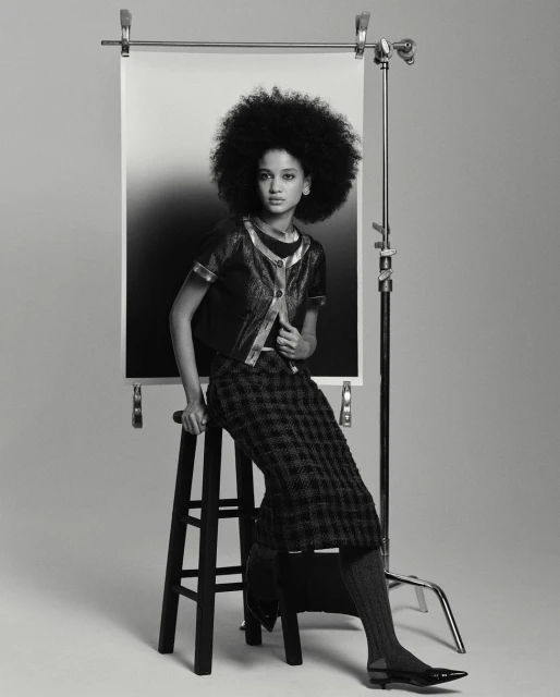 A woman with a large afro poses in a studio wearing a jacket and plaid skirt, sitting on a wooden stool with a gradient backdrop.