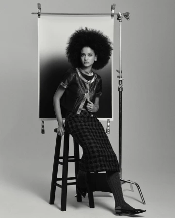 A woman with a large afro poses in a studio wearing a jacket and plaid skirt, sitting on a wooden stool with a gradient backdrop.