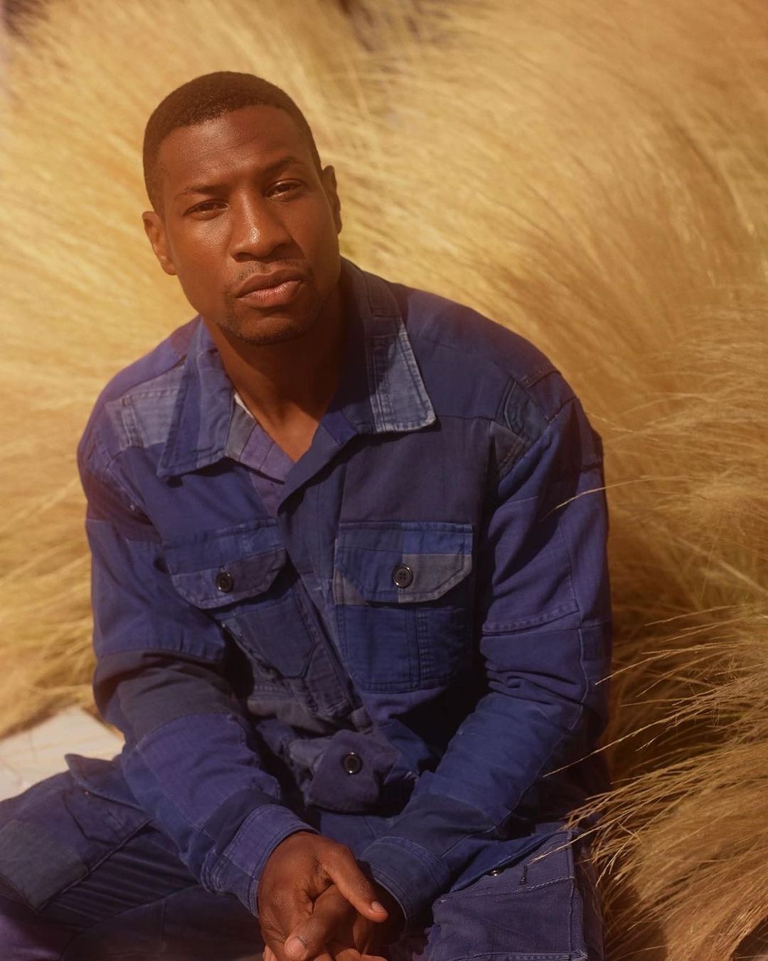 Portrait of a Black adult male in a blue workwear suit against a backdrop of soft golden wheat, seated indoors with eye-level camera angle.