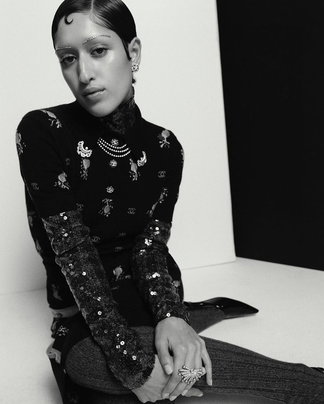 Black and white portrait of a female model with a turtleneck, adorned with jewelry and sequins, posing in a minimalist studio.