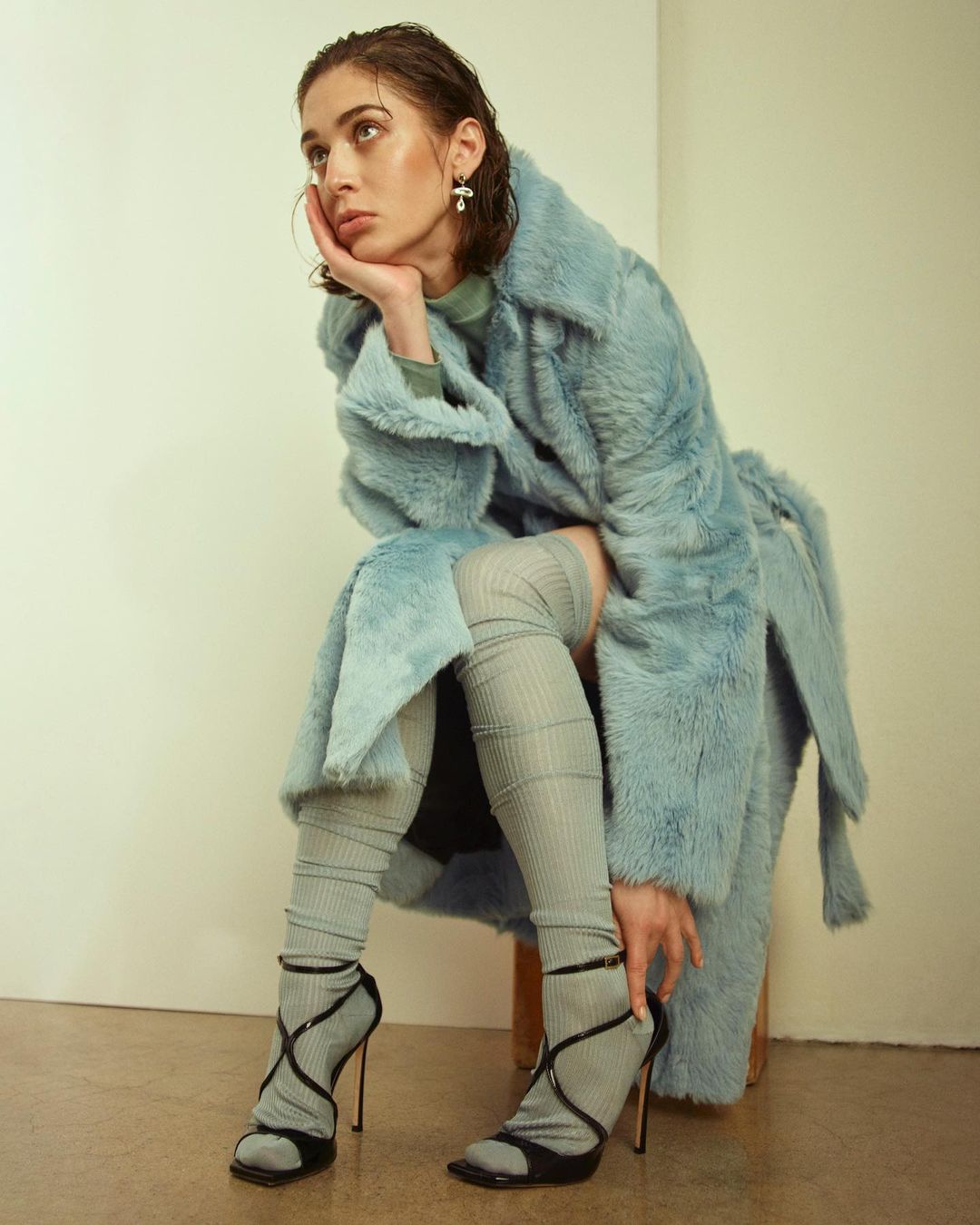 Woman in a light blue faux fur coat sits on a stool in a studio, wearing thigh-high socks and black high-heeled strappy shoes, looking off-camera.